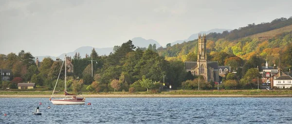 Güneşli bir günde yelkenli dolu bir yat. Arkasında tepeler, ormanlar ve kır evleri olan kayalık kıyılar. Rhu marinası, Clyde Firth, İskoçya, İngiltere