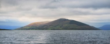 Bute 'un kayalık kıyılarının sudan panoramik görüntüsü. Tepeler ve dağlar arka planda. Karanlık fırtına gökyüzü. Bute Adası, Clyde Firth, İskoçya, İngiltere