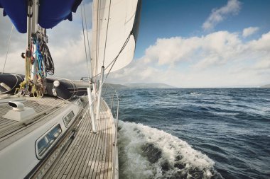 Bulutlu bir günde ahşap meşe güverteli, yelkenli, modern bir yat. Direğe ve yelkenlere bir manzara. Sudan kayalık nehir kıyılarının panoramik görüntüsü. Arka planda kır evleri, ağaçlar, tepeler ve dağlar var. Gare Gölü, Clyde Firth, S