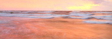 Gün batımında Baltık Denizi. Dramatik gökyüzü, mavi ve pembe parlayan bulutlar, yumuşak altın güneş ışığı. Dalgalar, su sıçratma. Deniz manzarası, bulut manzarası, doğa. Panoramik görünüm, uzun pozlama