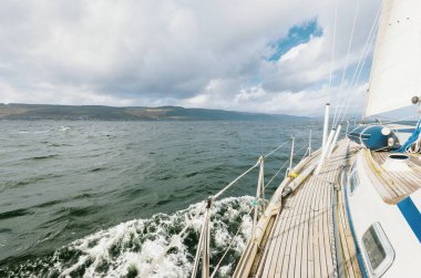 Teknelerle donatılmış modern bir yat. Tahta meşe güverteli. Clyde Nehri Firth 'ün kayalık kıyılarına doğru yol alıyorlar. Dramatik bir gökyüzü. İskoçya, İngiltere. Seyahat yerleri, eko turizm, tatiller, saf doğa.