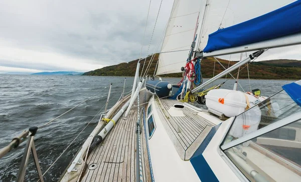 Sloop rigged modern yacht with wooden teak deck sailing near the rocky shores of Tarbert after the rain. Scotland, UK. Close-up view from the deck to the bow, mast and sails. Dramatic storm sky