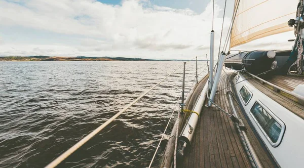 Sloop rigged modern yacht with wooden teak deck sailing near the rocky shores of Tarbert after the rain. Scotland, UK. Close-up view from the deck to the bow, mast and sails. Clear blue sky