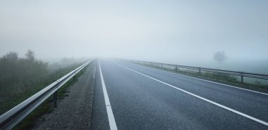 Gün doğumunda sisli bir havada boş otobanın tarlalar ve ormanlar arasındaki panoramik görüntüsü. Avrupa. Ulaşım, lojistik, seyahat, yolculuk, özgürlük, sürüş. Kırsal alan