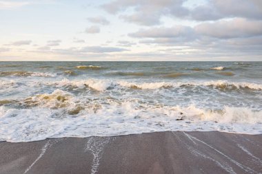 Gün batımında Baltık kıyıları (kum tepeleri). Yumuşak güneş ışığı, parlayan bulutlar, dalgalar ve su fışkıran berrak gökyüzü. Idyllic deniz manzarası. Liepaja, Letonya, Avrupa. Sıcak kış havası, iklim değişikliği, doğa