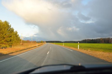 Gün batımında tarla ve orman boyunca asfalt yol (karayolu). Arabadan görüntü. Turizm, tatiller, uzak yerler, lojistik kavramlar