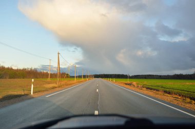 Gün batımında tarla ve orman boyunca asfalt yol (karayolu). Arabadan görüntü. Turizm, tatiller, uzak yerler, lojistik kavramlar