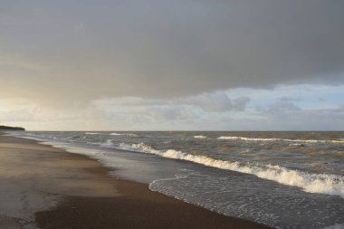 Gün batımında Baltık kıyıları (kum tepeleri). Yumuşak güneş ışığı, parlayan bulutlar, dalgalar ve su fışkıran berrak gökyüzü. Idyllic deniz manzarası. Liepaja, Letonya, Avrupa. Sıcak kış havası, iklim değişikliği, doğa