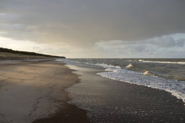 Gün batımında Baltık kıyıları (kum tepeleri). Yumuşak güneş ışığı, parlayan bulutlar, dalgalar ve su fışkıran berrak gökyüzü. Idyllic deniz manzarası. Liepaja, Letonya, Avrupa. Sıcak kış havası, iklim değişikliği, doğa