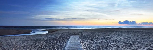 Starry sky with noctilucent clouds above the Baltic sea shore at summer solstice night. An empty wooden pathway. Golden flowing sunlight. Long exposure. Fantastic cloudscape. Latvia