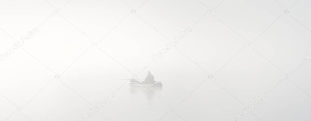 Fisherman in an inflatable boat, lake in a thick white morning fog. Atmospheric monochrome black and white landscape. Fishing, hobby sport, recreation, ecotourism, silence, loneliness concepts