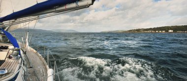 Bulutlu bir günde ahşap meşe güverteli, yelkenli, modern bir yat. Direğe ve yelkenlere bir manzara. Sudan kayalık nehir kıyılarının panoramik görüntüsü. Arka planda kır evleri, ağaçlar, tepeler ve dağlar var. Gare Gölü, Clyde Firth, S