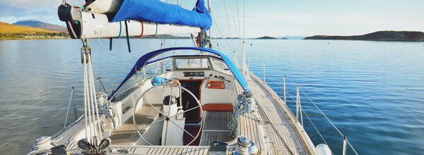 Sloop rigged modern yacht with wooden teak deck sailing near the shore of Jura island on a sunny day. Cloudy blue sky. Reflections on the water. Inner Hebrides, Scotland, UK