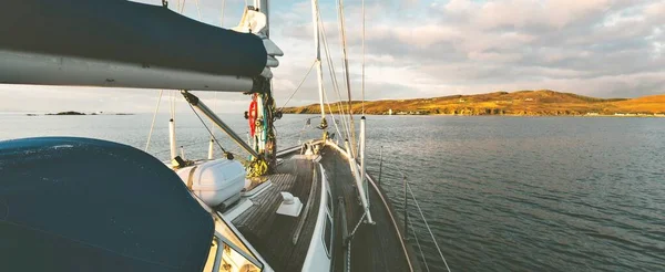 Sloop rigged modern yacht with wooden teak deck sailing near the shore of Isle of Islay at sunset. Inner Hebrides, Scotland, UK. Sport and recreation, travel destinations, landmarks, transportation