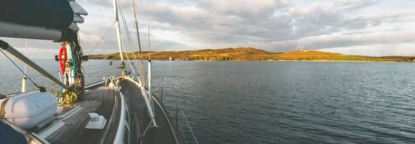 Sloop rigged modern yacht with wooden teak deck sailing near the shore of Isle of Islay at sunset. Inner Hebrides, Scotland, UK. Sport and recreation, travel destinations, landmarks, transportation