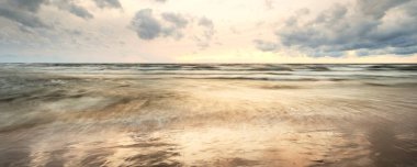 Fırtınadan sonra Baltık kıyıları. Dramatik günbatımı gökyüzü, karanlık parlayan bulutlar, altın güneş ışığı. Dalgalar, su sıçratma. Resimli manzara, deniz manzarası, bulut manzarası, doğa. Panoramik görünüm, uzun pozlama