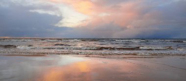 Kumlu bir sahilden Baltık Denizi 'nin panoramik manzarası. Açık günbatımı gökyüzü, parlak pembe bulutlar, yumuşak ışık. Suyun simetri yansımaları, doğal ayna. Hava, kış, iklim değişikliği, doğa