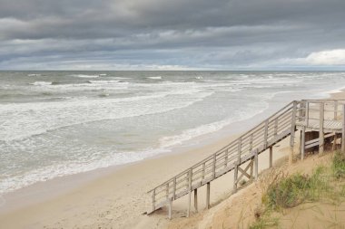 Fırtınadan sonra Baltık kıyısı (plaj). Modern tahta kaldırım (tahta kaldırım). Kum tepeleri. Dramatik gökyüzü, dalgalar ve su sıçramaları. Doğa, ekoloji, eko-turizm, yürüyüş, eğlence kavramları