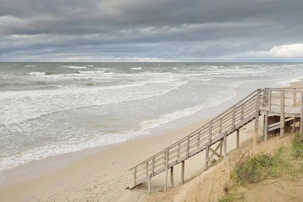 Fırtınadan sonra Baltık kıyısı (plaj). Modern tahta kaldırım (tahta kaldırım). Kum tepeleri. Dramatik gökyüzü, dalgalar ve su sıçramaları. Doğa, ekoloji, eko-turizm, yürüyüş, eğlence kavramları
