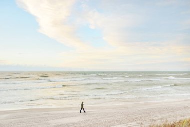 Gün batımında Baltık kıyısı. Kumsal, kum tepeleri. Açık günbatımı gökyüzü, parlayan bulutlar. Cep telefonuyla yürüyen yalnız insanlar. Yerel seyahat, eko turizm, zihinsel sağlık, sosyal sorunlar ve uzaklık temaları