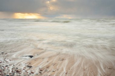 Gün batımında Baltık Denizi. Dramatik gökyüzü, mavi ve pembe parlayan bulutlar, altın güneş ışığı. Dalgalar, su sıçratma. Resimli manzara, deniz manzarası, doğa. Panoramik görünüm, uzun pozlama