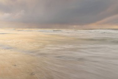 Gün batımında Baltık Denizi. Dramatik gökyüzü, mavi ve pembe parlayan bulutlar, altın güneş ışığı. Dalgalar, su sıçratma. Resimli manzara, deniz manzarası, doğa. Panoramik görünüm, uzun pozlama