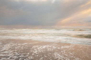 Gün batımında Baltık Denizi. Dramatik gökyüzü, mavi ve pembe parlayan bulutlar, altın güneş ışığı. Dalgalar, su sıçratma. Resimli manzara, deniz manzarası, doğa. Panoramik görünüm, uzun pozlama