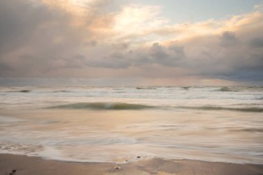 Gün batımında Baltık Denizi. Dramatik gökyüzü, mavi ve pembe parlayan bulutlar, altın güneş ışığı. Dalgalar, su sıçratma. Resimli manzara, deniz manzarası, doğa. Panoramik görünüm, uzun pozlama