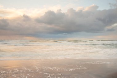 Gün batımında Baltık Denizi. Dramatik gökyüzü, mavi ve pembe parlayan bulutlar, altın güneş ışığı. Dalgalar, su sıçratma. Resimli manzara, deniz manzarası, doğa. Panoramik görünüm, uzun pozlama