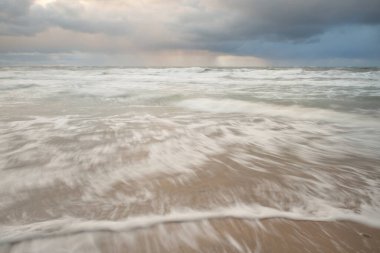Gün batımında Baltık Denizi. Dramatik gökyüzü, mavi ve pembe parlayan bulutlar, altın güneş ışığı. Dalgalar, su sıçratma. Resimli manzara, deniz manzarası, doğa. Panoramik görünüm, uzun pozlama