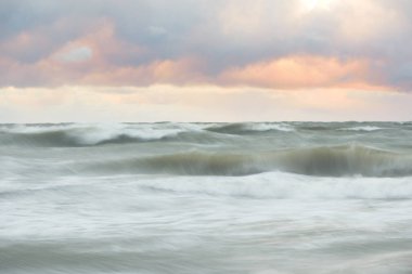 Gün batımında Baltık Denizi. Dramatik gökyüzü, mavi ve pembe parlayan bulutlar, altın güneş ışığı. Dalgalar, su sıçratma. Resimli manzara, deniz manzarası, doğa. Panoramik görünüm, uzun pozlama