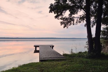 Forest lake at sunset. Wooden pier. Soft sunlight, glowing clouds, symmetry reflections in a crystal clear water. Idyllic landscape. Panoramic view. Nature, ecology, ecotourism. Peace and joy concepts