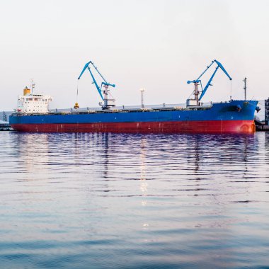 Liman terminalinde büyük bir kargo gemisi yükleniyor, arka planda vinçler var. Yük taşımacılığı, deniz taşımacılığı, lojistik, küresel iletişim, dünya çapında taşımacılık, ekonomi, ticaret, sanayi