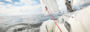 Açık bir günde açık denizde yelken açan beyaz yelkenli bir yat. Güverteden pruvaya bir manzara. Kümülüs bulutları. Ulaşım, seyahat, gezi, spor, eğlence, boş zaman aktivitesi, yarış, yelkenli yarışı