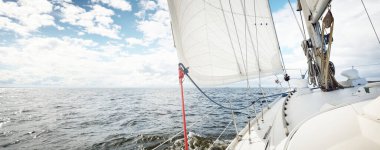 Açık bir günde açık denizde yelken açan beyaz yelkenli bir yat. Güverteden pruvaya bir manzara. Kümülüs bulutları. Ulaşım, seyahat, gezi, spor, eğlence, boş zaman aktivitesi, yarış, yelkenli yarışı