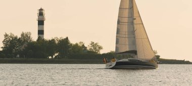 Baltık Denizi 'nde yelkenli bir yat. Deniz fenerine yakın çekim. Fırtınadan sonra gün batımı gökyüzü açık. Ulaşım, seyahat, gezi, spor, eğlence, boş zaman aktivitesi, yarış, yelkenli yarışı