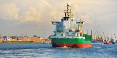 Büyük yeşil yük gemisi gün batımında kargo terminaline varacak. Arka planda gemiler ve vinçler var. Dramatik bulut manzarası. Yük taşımacılığı, küresel iletişim, endüstri, ekonomi, lojistik