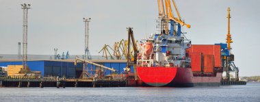 Liman terminalinde büyük bir kargo gemisi yükleniyor, arka planda vinçler var. Yük taşımacılığı, lojistik, küresel iletişim, ekonomi, ticaret, sanayi, özel ekipman