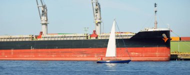 Baltık Denizi 'nde yelkenli bir yat. Kargo gemisi arka plandaki iskele terminaline yükleniyor. Ulaşım, seyahat, gezinti, spor, eğlence, eğlence etkinliği, iş, ekonomi, sanayi