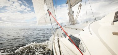 Açık bir günde açık denizde yelken açan beyaz yelkenli bir yat. Güverteden pruvaya bir manzara. Kümülüs bulutları. Ulaşım, seyahat, gezi, spor, eğlence, boş zaman aktivitesi, yarış, yelkenli yarışı