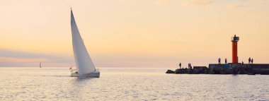 Baltık Denizi 'nde beyaz yelkenli yelkenli. Deniz feneri, yürüyen insanlar. Fırtınadan sonra gün batımı gökyüzü açık. Ulaşım, seyahat, gezi, spor, eğlence, boş zaman aktivitesi, yelkenli yarışı