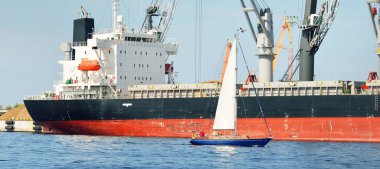 Baltık Denizi 'nde yelkenli bir yat. Kargo gemisi arka plandaki iskele terminaline yükleniyor. Ulaşım, seyahat, gezinti, spor, eğlence, eğlence etkinliği, iş, ekonomi, sanayi