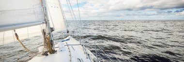 Açık bir günde açık denizde yelken açan beyaz yelkenli bir yat. Güverteden pruvaya bir manzara. Kümülüs bulutları. Ulaşım, seyahat, gezi, spor, eğlence, boş zaman aktivitesi, yarış, yelkenli yarışı