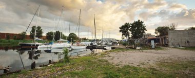 Günbatımında yat limanında demirlemiş zarif ve modern yelkenliler. Dramatik gökyüzü, parlayan altın bulutlar. Tatiller, ulaşım, spor, eğlence, turizm, seyahat, liman