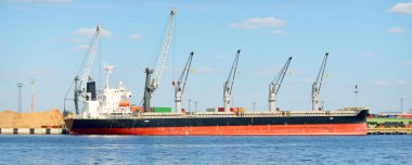 Liman terminalinde büyük bir kargo gemisi yükleniyor, arka planda vinçler var. Yük taşımacılığı, lojistik, küresel iletişim, ekonomi, ticaret, sanayi, özel ekipman