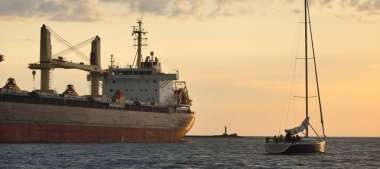 Büyük kargo gemisi (büyük yük gemisi, 179.99 metre uzunluğunda) ve günbatımında Baltık Denizi 'nde yelkenli yelkenli. Yük taşımacılığı, küresel iletişim, ekonomi, seyahat, eğlence. spor