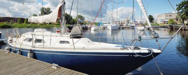 Güneşli bir yaz gününde mavi şalopalı bir yat rıhtıma demirli. Arka planda yelkenliler var. Baltık Denizi, Avrupa. Tatiller, ulaşım, spor, eğlence, turizm, seyahat, balıkçılık, liman hizmeti