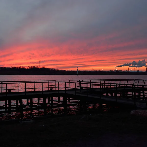 Gün batımında nehir. Dramatik gökyüzü, parlayan kırmızı bulutlar. Arka planda port terminali. Kış manzarası kavramı. Doğa, ekoloji, çevresel hasar, kirlilik, iklim değişikliği