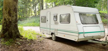 Beyaz karavan karavanı ve muhteşem ağaçların altındaki kırsal yolda park edilmiş bir araba. Orman kamp alanı. Avrupa. Yaşam tarzı, özgürlük, seyahat, eko-turizm, yolculuk, dinlenme, ulaşım, karavan, karavan