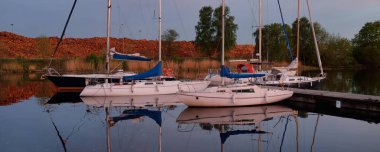 Günbatımında yat limanında demirlemiş zarif ve modern yelkenliler. Dramatik gökyüzü, parlayan altın bulutlar. Tatiller, ulaşım, spor, eğlence, turizm, seyahat, liman